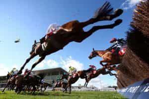Horses clear a fence at Cheltenham Racecourse with a camera shooting the action from below.