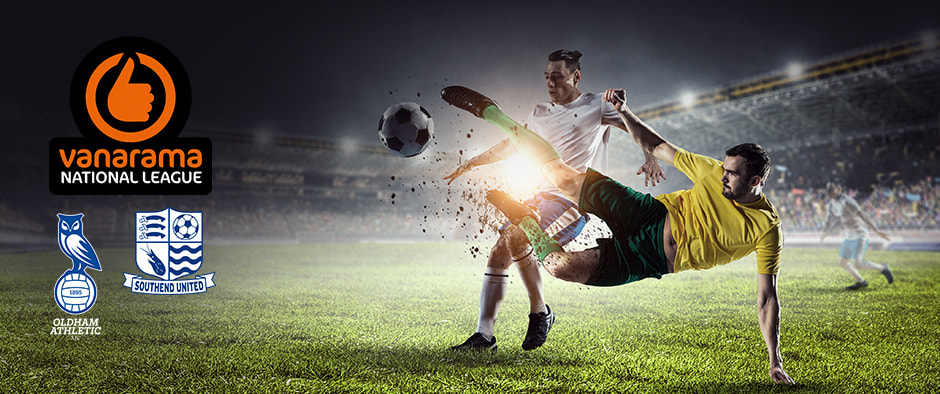 A dramatic action shot on a soccer field under stadium lights, showing two male football players in mid-tackle competing for the ball. One player, in a white jersey and blue shorts, clashes with another in a yellow jersey and green shorts, with turf flying from the impact. On the left side of the image are the Vanarama National League logo and the crests of Oldham Athletic and Southend United, highlighting a match between the two teams. The background features a packed stadium and a blurred player in motion, capturing the intensity of the game.