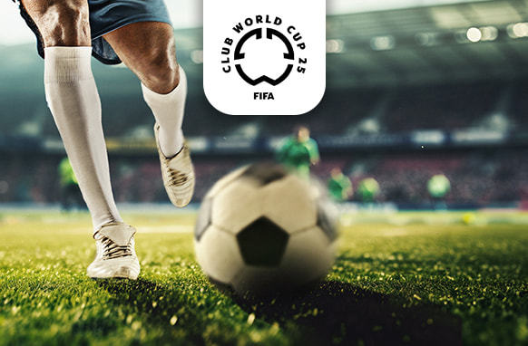 A close-up action image of a football player on the verge of kicking a black-and-white football on a sunlit grass pitch, inside a large stadium. The player's lower body is visible, wearing blue shorts, white socks, and white football boots. In the background, other players can be seen as blurred figures. The FIFA Club World Cup 25 logo is prominently displayed above, indicating an important football event.