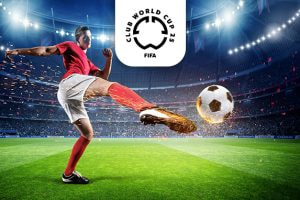 The image captures a football player wearing a red shirt and shorts energetically kicking a football inside a brightly lit stadium. Sparks fly as the ball is struck, emphasising the power and motion of the kick. Above the scene is the logo for the FIFA Club World Cup 2025, positioned prominently. The stadium is filled with an electrifying atmosphere, highlighting the intensity of the match.