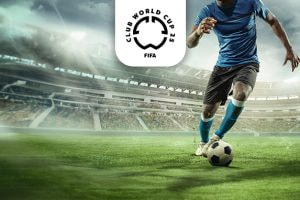 The image features a football player in a blue shirt and shorts controlling a football on a grass field within a large stadium. The stadium's architecture is visible, with empty stands in the background. Above the player, the FIFA Club World Cup 2025 logo is centrally positioned. The setting conveys a sense of focus and action on the field.