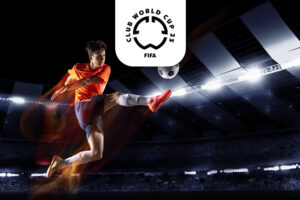 The image features a football player in an orange shirt and blue shorts executing a powerful mid-air kick inside a brightly lit stadium at night. The ball is seen in motion, highlighting the player's athleticism and focus. Above the scene, the FIFA Club World Cup 2025 logo is centrally placed. The stadium's backdrop adds to the dynamic and exciting atmosphere of the moment.