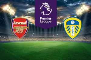 The image shows the logos of two Premier League football clubs, Arsenal and Leeds United, displayed on a football pitch background. The Arsenal logo is on the left side, featuring a red shield with a cannon and the text "Arsenal." The Leeds United logo is on the right side, featuring a shield with yellow and blue stripes, a white rose at the top, and the text "LUFC." In the centre, there is the Premier League logo, consisting of a lion's head above the text "Premier League." The background depicts a lit football stadium with a dramatic, cloudy sky above, creating an intense and competitive atmosphere.