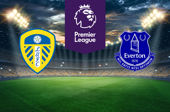 The image shows the logos of two Premier League football clubs, Leeds United and Everton, displayed on a football pitch background. The Leeds United logo is on the left side, featuring a shield with a white rose on top and blue and yellow stripes, along with the text "LUFC." The Everton logo is on the right side, featuring a royal blue crest with a tower, laurels, and the text "Everton 1878" and "Nil Satis Nisi Optimum." In the centre, there is the Premier League logo, consisting of a lion's head above the text "Premier League." The background depicts a lit football stadium with a dramatic, cloudy sky above.