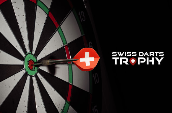 The image features a close-up of a dartboard with a dart successfully hitting the bullseye. The dart's flight is adorned with a red design featuring a white cross, reminiscent of the Swiss flag. To the right of the dartboard, the text "Swiss Darts Trophy" is displayed, with the word "Trophy" stylised to include a small red diamond with a white cross in place of the letter "O" in "Trophy," echoing the Swiss national flag. The overall visual composition emphasises precision and national pride, creating a focused and compelling atmosphere for the Swiss Darts Trophy event.