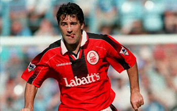 Dean Saunders in action wearing a red and black football kit with "Labatt's" on the front. He appears to be running on the field with a focused expression. The background shows blurred stadium seats and spectators.