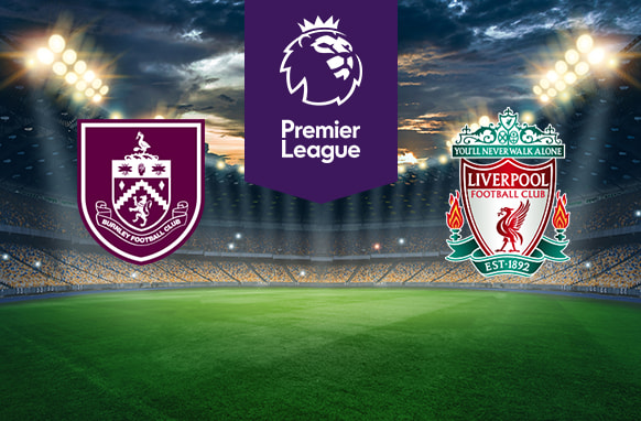 The image shows the logos of two football clubs, Burnley and Liverpool, displayed on a football pitch. The Burnley logo is on the left, featuring a maroon and white crest with various symbols and the text "Burnley Football Club." The Liverpool logo is on the right, featuring a green, red, and white crest with a liver bird and the text "Liverpool Football Club" along with the motto "You'll Never Walk Alone" and the establishment year "EST 1892." In the centre above the pitch is the Premier League logo, which includes a lion's head and the text "Premier League" below it on a purple background. The background depicts a stadium with dramatic lighting, creating an energetic and intense atmosphere.
