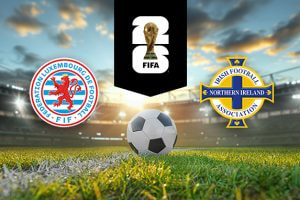 The image shows the logos of two national football associations, the Luxembourg Football Federation and the Irish Football Association (Northern Ireland), displayed on a football pitch background with a football placed in the centre. The Luxembourg logo is on the left side, featuring a red lion within a circular design, with the words "Fédération Luxembourgeoise de Football" and "FLF" written around the circle in red and blue. The Northern Ireland logo is on the right side, featuring a blue and gold cross with the text "Irish Football Association" and "Northern Ireland" integrated into the design. In the centre, above the football, there is the FIFA logo, consisting of the FIFA World Cup Trophy and the text "FIFA" below it on a black background. The backdrop depicts a football stadium with a dramatic sky, either during sunrise or sunset, creating a dynamic and compelling atmosphere.