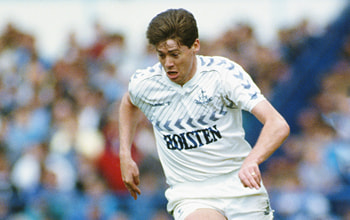 The image features a football player in action during a match. He is wearing a white football kit with a chevron pattern on the shoulders and the text "HOLSTEN" across the front. The player is mid-run, leaning forward, and appears focused on the game. His hair is short and neatly styled, and he has a determined expression on his face. The background shows a blurred crowd of spectators, indicating the match's lively atmosphere.