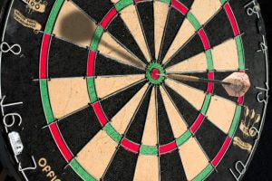 The image captures a close-up view of a dartboard, focusing on the concentric rings and numbered sections that represent different point zones. A dart is embedded in the board, striking the bullseye precisely at the center, highlighting a perfect throw. The dartboard is divided into alternating black and cream segments, with green and red outer and inner bullseyes. The numbering around the circumference of the dartboard designates the scoring areas, ensuring a fair game. The scene is well-lit, emphasizing the accuracy and precision required in the game of darts.