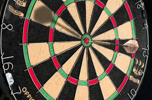 The image captures a close-up view of a dartboard, focusing on the concentric rings and numbered sections that represent different point zones. A dart is embedded in the board, striking the bullseye precisely at the center, highlighting a perfect throw. The dartboard is divided into alternating black and cream segments, with green and red outer and inner bullseyes. The numbering around the circumference of the dartboard designates the scoring areas, ensuring a fair game. The scene is well-lit, emphasizing the accuracy and precision required in the game of darts.