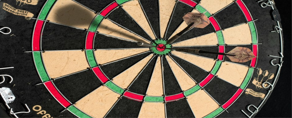 The image presents a detailed view of a dartboard, prominently displaying its various scoring segments and patterns. The board features alternating black and beige sections, with green and red areas marking the outer and inner bullseyes. Two darts are lodged in the board, one hitting the bullseye and the others close to it, demonstrating impressive precision in the throws. The surrounding numbers along the edge designate different point zones, guiding the scoring rules. The board displays signs of extensive use, characterised by small marks and wear, emphasising the competitive spirit of the game. The lighting in the image enhances the clarity of the board's details and the accuracy of the dart placements.